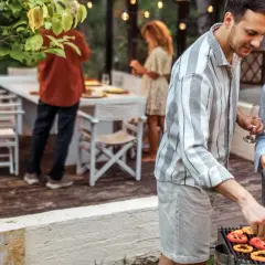 Un homme et une femme préparent un barbecue. Des poivrons, du maïs et des saucisses cuisent sur le gril. Un verre de vin blanc est à portée de main.