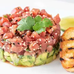 Délicieux tartare de bar frais, garni de sésame et de coriandre, servi avec des toasts grillés et une tranche de citron vert.
