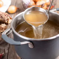 Une louche verse un bouillon doré dans une casserole, sur un fond brun. Des queues de bœuf et des oignons sont disposés autour.