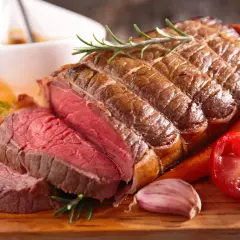 Un délicieux roti de boeuf au four, saignant et tendre, servi avec des carottes, une tomate et de l'ail sur une planche en bois.