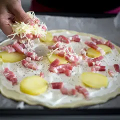 Une main saupoudre du fromage râpé sur une pizza base crème garnie de lardons et de fines rondelles de pommes de terre.