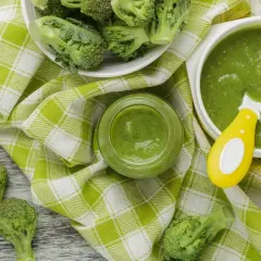 Un bol et un pot en verre remplis de purée de brocolis, avec des fleurettes de brocolis fraîches disposées autour. Une cuillère jaune est dans le bol.