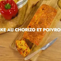 Un cake au chorizo et poivrons, coupé en tranche, sur une planche en bois. Un poivron rouge est posé à côté.