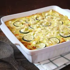 Un plat blanc rectangulaire contient un gratin de courgettes moelleux, garni de rondelles de courgettes et de morceaux de fromage. Une cuillère en bois repose à côté.