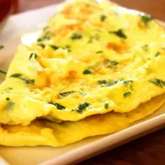 Une omelette au fromage, dorée et moelleuse, parsemée d'herbes fraîches, servie sur une assiette blanche.