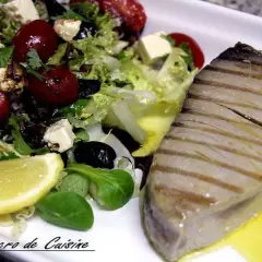 Un steak de thon au barbecue, grillé à la perfection, servi avec une salade fraîche composée de laitue, tomates cerises, olives noires, feta et un quartier de citron.