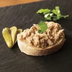 Tranche de pain garnie de rillettes de porc, accompagnée de cornichons et d'une feuille de persil.