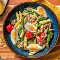 Salade de penne colorée avec haricots verts, tomates cerises, thon et œufs durs, servie dans un plat bleu.