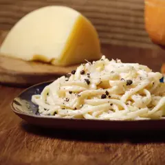 Un plat de pâtes crémeux, garni de poivre noir concassé, évoque le goût authentique du cacio e pepe. Un morceau de fromage pecorino est visible en arrière-plan.
