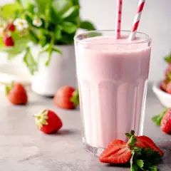 Un délicieux milkshake fraise crémeux, garni de pailles rayées, entouré de fraises fraîches et d'un plant de fraises en arrière-plan.