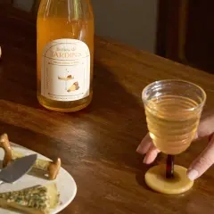 Une main saisit un verre de ce qui pourrait être le meilleur vin blanc moelleux, à côté d'une bouteille "Bulles de Jardins" et de fromages.