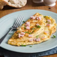 Une délicieuse omelette jambon fromage, garnie de ciboulette et de poivre noir, prête à être dégustée avec une tranche de pain croustillant.