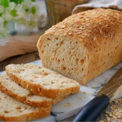 Un pain complet maison, doré et garni de flocons d'avoine, est tranché sur une planche en marbre. Des épis de blé et un couteau complètent la scène.