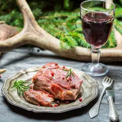 Un plat de chevreuil rôti, garni de romarin et de baies roses, accompagné d'un verre de vin rouge. La question est : quel vin avec du chevreuil ?