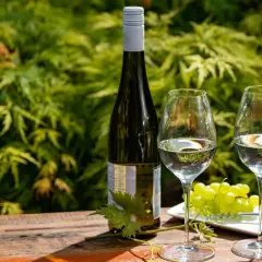Bouteille de riesling avec deux verres, une grappe de raisin et un tire-bouchon sur une table en bois, devant un feuillage verdoyant.