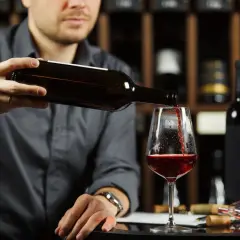 Un homme verse un vin rouge profond dans un verre, une cave à vin en arrière-plan. Quel vin rouge savoureux !
