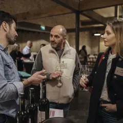 Trois personnes discutent autour d'une table de dégustation. L'une d'elles tient un verre de vin, peut-être un rosé de la terre du midi.