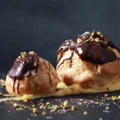 Deux profiteroles au chocolat, garnis de crème pâtissière et saupoudrés de pistaches concassées, sur un fond sombre.
