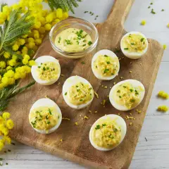Des œufs mimosa garnis d'une crème onctueuse et de ciboulette, disposés sur une planche en bois, avec des fleurs de mimosa en arrière-plan.