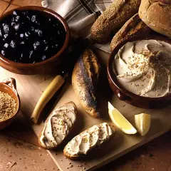 Un bol de purée de pois chiche crémeuse, garnie de graines, avec du pain, des olives noires et des quartiers de citron.