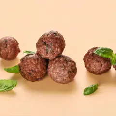 Cinq boulettes de viande dorées, garnies de feuilles de basilic frais, sur fond beige.