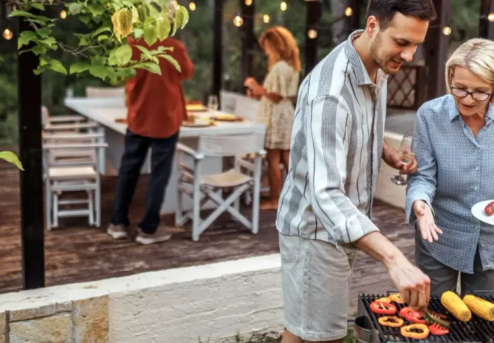 Un homme et une femme préparent un barbecue. Des poivrons, du maïs et des saucisses cuisent sur le gril. Un verre de vin blanc est à portée de main.