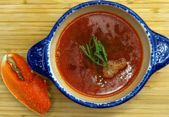 Une délicieuse bisque de homard, garnie d'herbes fraîches, est servie dans un bol bleu et blanc, accompagnée d'une pince de homard sur une planche en bois.