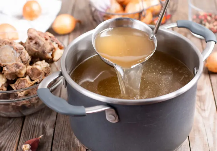 Une louche verse un bouillon doré dans une casserole, sur un fond brun. Des queues de bœuf et des oignons sont disposés autour.