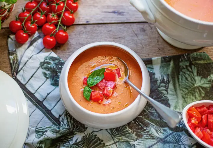 Un bol de soupe à la tomate fraîchement préparée, garnie de dés de tomates et de basilic. Des tomates cerises fraîches sont disposées à côté.