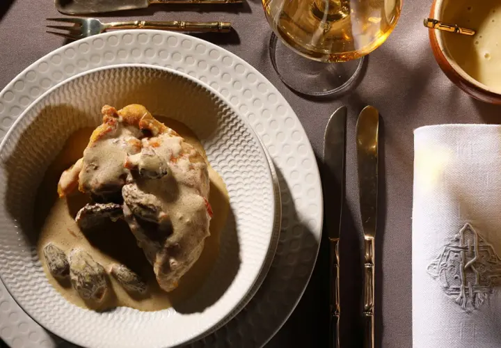 Un plat de poulet au vin jaune, crémeux et garni de morilles, servi sur une assiette texturée. Une table dressée avec élégance.