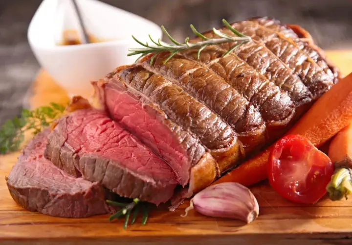 Un délicieux roti de boeuf au four, saignant et tendre, servi avec des carottes, une tomate et de l'ail sur une planche en bois.