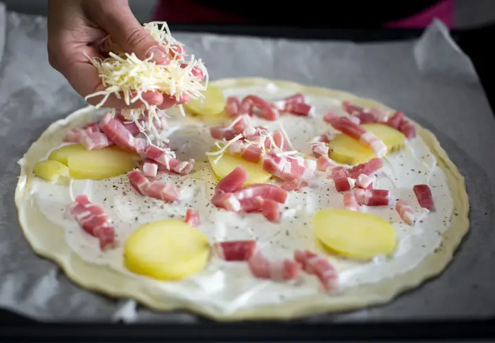 Une main saupoudre du fromage râpé sur une pizza base crème garnie de lardons et de fines rondelles de pommes de terre.