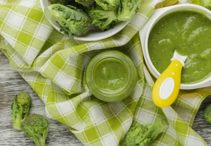Un bol et un pot en verre remplis de purée de brocolis, avec des fleurettes de brocolis fraîches disposées autour. Une cuillère jaune est dans le bol.
