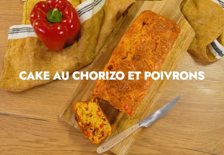 Un cake au chorizo et poivrons, coupé en tranche, sur une planche en bois. Un poivron rouge est posé à côté.