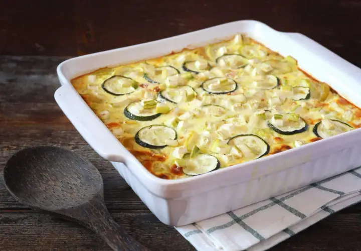 Un plat blanc rectangulaire contient un gratin de courgettes moelleux, garni de rondelles de courgettes et de morceaux de fromage. Une cuillère en bois repose à côté.