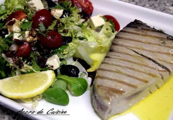 Un steak de thon au barbecue, grillé à la perfection, servi avec une salade fraîche composée de laitue, tomates cerises, olives noires, feta et un quartier de citron.