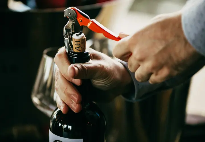 Un homme ouvre une bouteille de vin rouge avec un tire-bouchon. La scène évoque le plaisir de la dégustation et la conservation du vin.