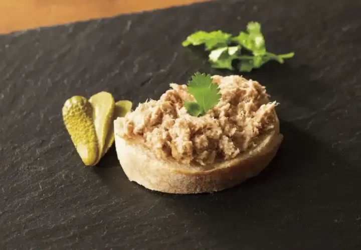 Tranche de pain garnie de rillettes de porc, accompagnée de cornichons et d'une feuille de persil.