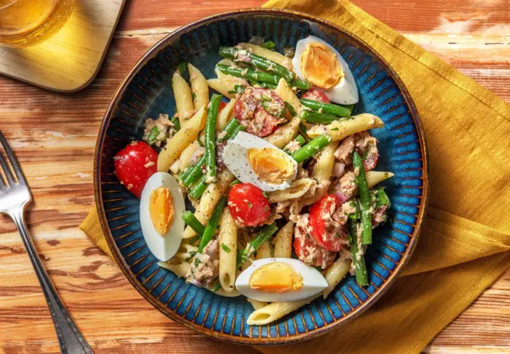 Salade de penne colorée avec haricots verts, tomates cerises, thon et œufs durs, servie dans un plat bleu.