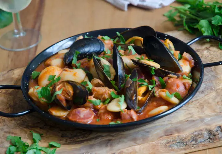 Un plat de moules au chorizo, garni de persil frais, servi dans une poêle noire sur une planche en bois.