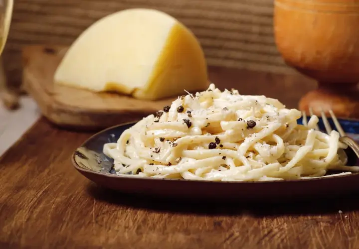 Un plat de pâtes crémeux, garni de poivre noir concassé, évoque le goût authentique du cacio e pepe. Un morceau de fromage pecorino est visible en arrière-plan.