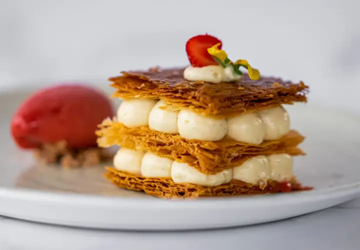 Un délicieux millefeuille croustillant, garni de crème onctueuse et d'une fraise, servi avec une boule de sorbet rouge.