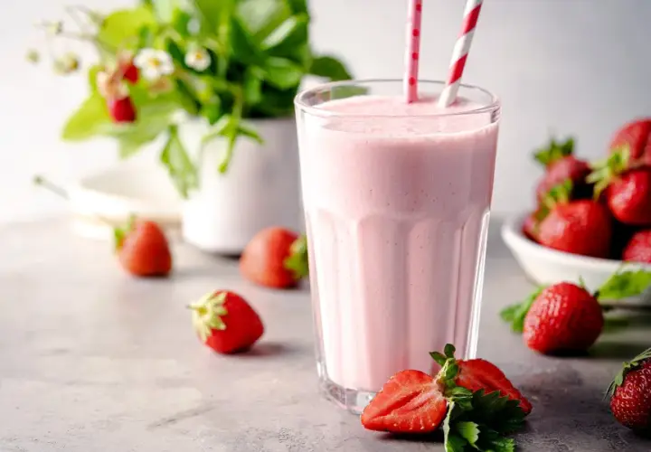 Un délicieux milkshake fraise crémeux, garni de pailles rayées, entouré de fraises fraîches et d'un plant de fraises en arrière-plan.