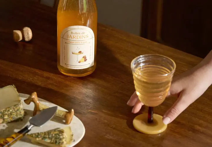 Une main saisit un verre de ce qui pourrait être le meilleur vin blanc moelleux, à côté d'une bouteille "Bulles de Jardins" et de fromages.
