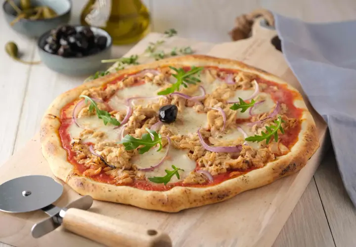 Une délicieuse pizza au thon, garnie d'oignons rouges, de roquette et d'une olive noire, prête à être découpée.