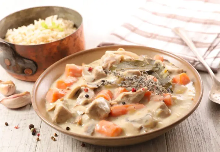 Un plat de blanquette de veau crémeuse avec des carottes et des champignons, servi avec du riz. Un verre de vin rouge est suggéré pour accompagner ce délice.