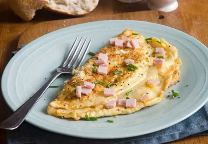 Une délicieuse omelette jambon fromage, garnie de ciboulette et de poivre noir, prête à être dégustée avec une tranche de pain croustillant.