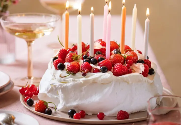 Pavlova fruit rouge, ornée de fraises, framboises et myrtilles, avec des bougies allumées pour une fête gourmande.