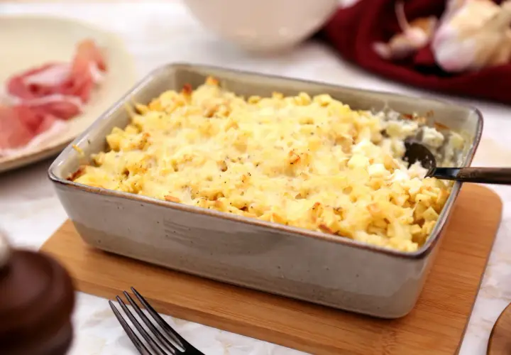 Un plat de gratin de crozet doré et gratiné, prêt à être dégusté. Une assiette de jambon cru est visible à gauche.
