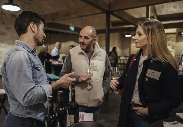 Trois personnes discutent autour d'une table de dégustation. L'une d'elles tient un verre de vin, peut-être un rosé de la terre du midi.